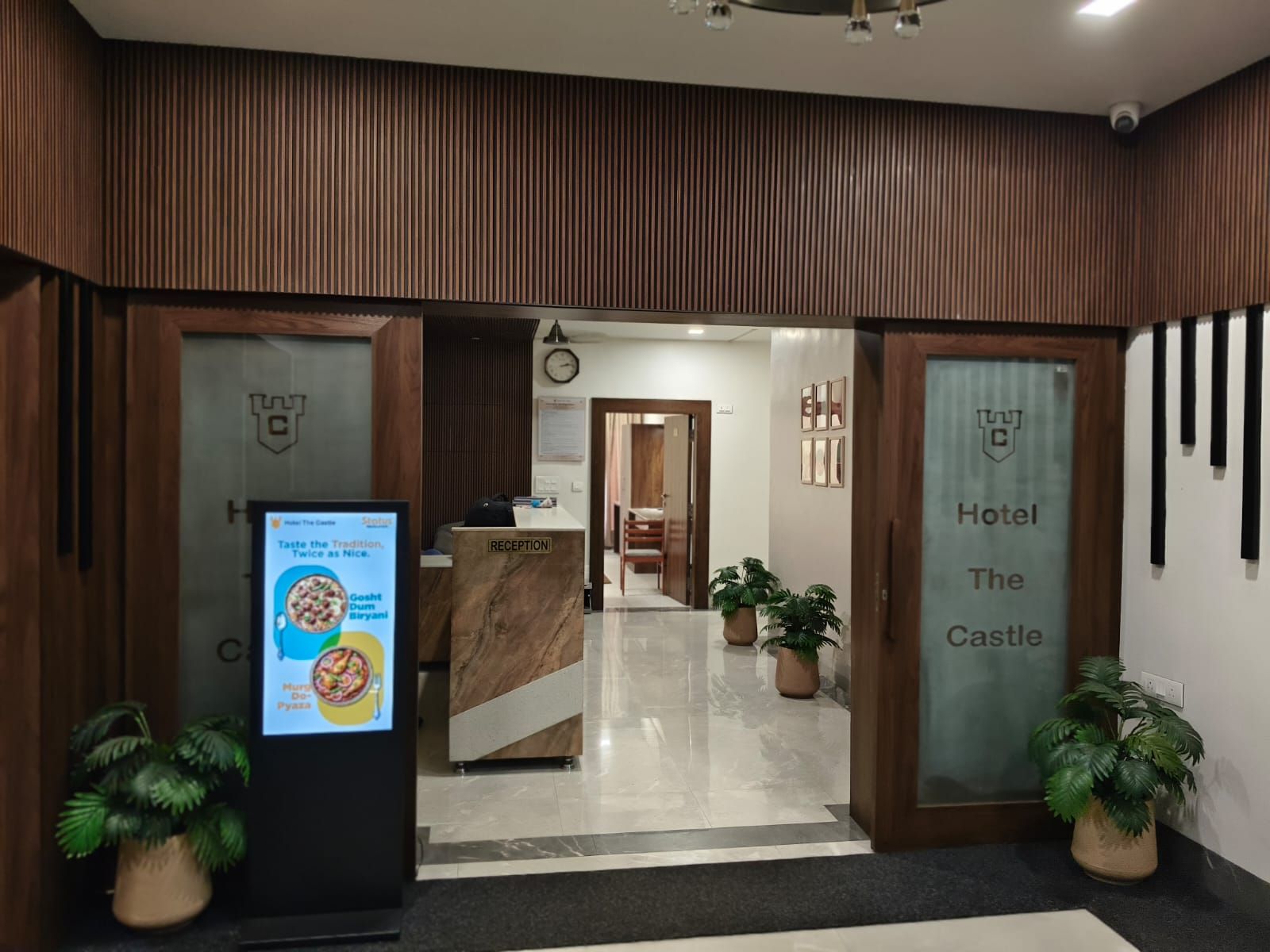 A hotel reception area with a reception desk, a clock, and potted plants.