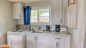 A compact kitchen with white cabinets, a stove, a sink, and a window with blue curtains. - Cozy Lake Escape: 2-Bed with Bunk, Sleeps 4, Full Kitchen & AC (Perth Road)