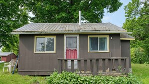 A small, single-story cabin with a red door, a front porch, and a metal roof. - Cozy Lake Escape: 2-Bed with Bunk, Sleeps 4, Full Kitchen & AC (Perth Road)