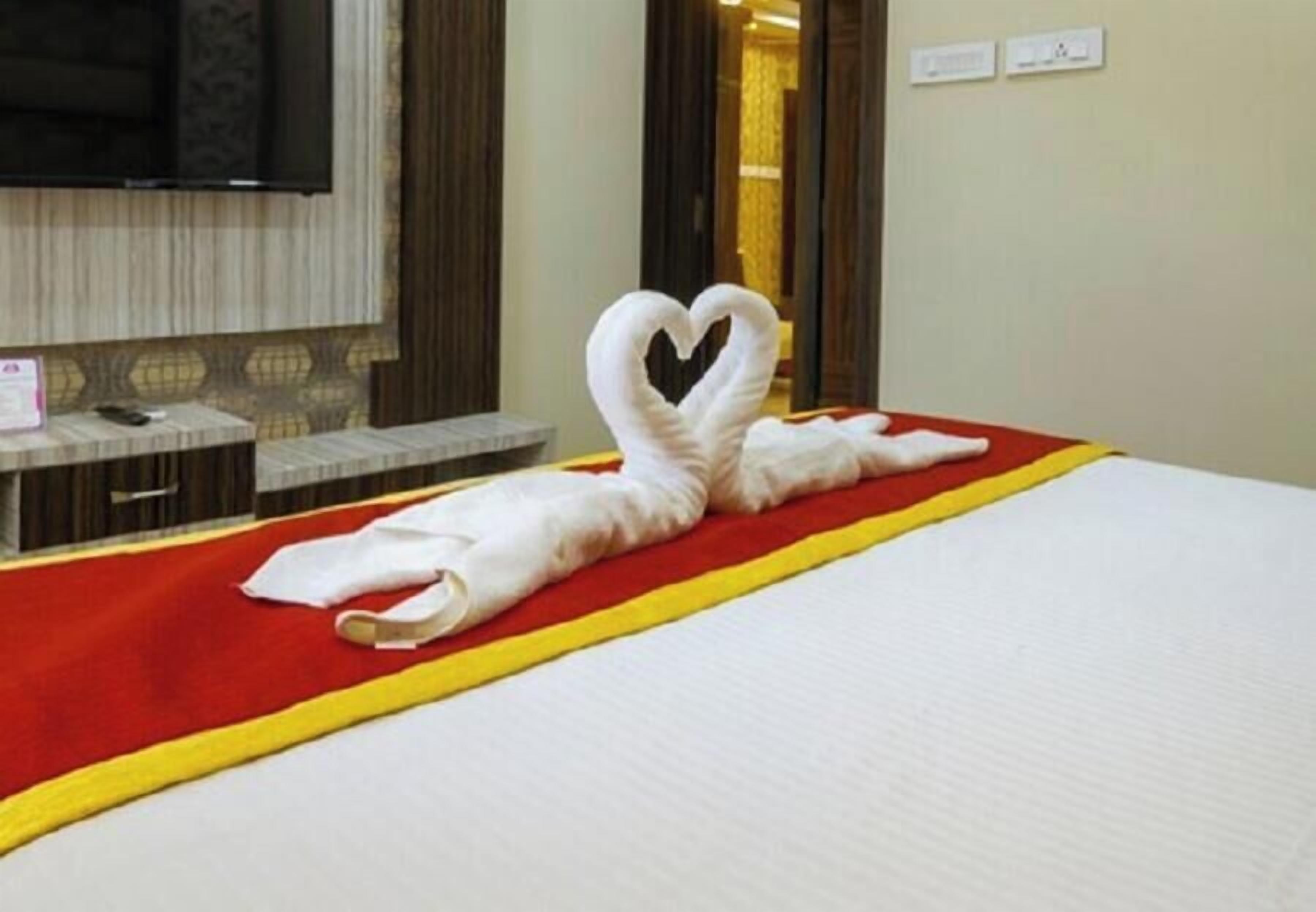 A hotel room with a bed, a heart-shaped towel, a red and yellow bedspread, a television, and a mirror.