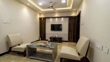 Un salon moderne avec une table basse en verre, deux fauteuils, un ventilateur de plafond, un éclairage encastré et un téléviseur à écran plat fixé au mur.