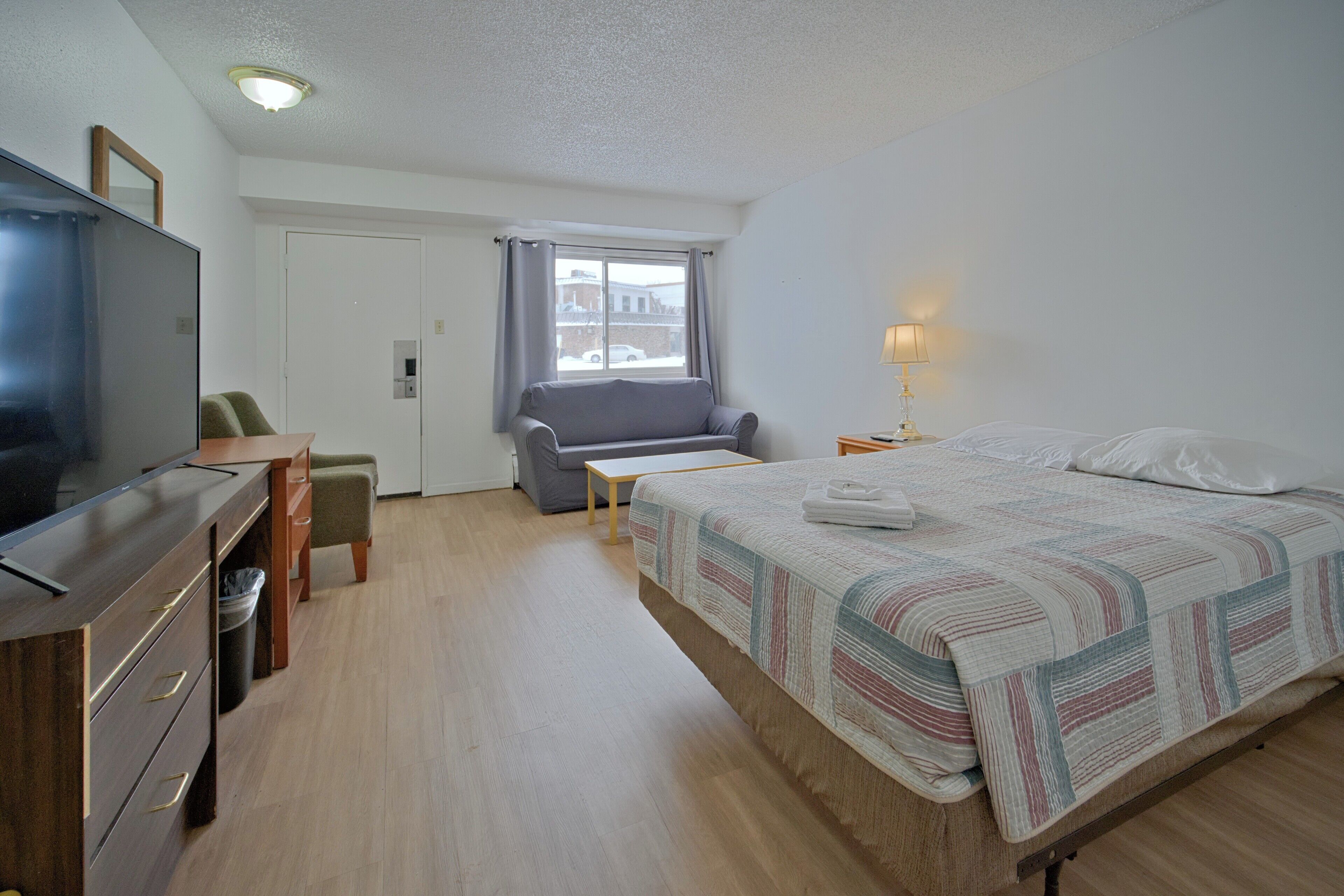A hotel room with a bed, a TV, a desk, a chair, a sofa, and a window with curtains.