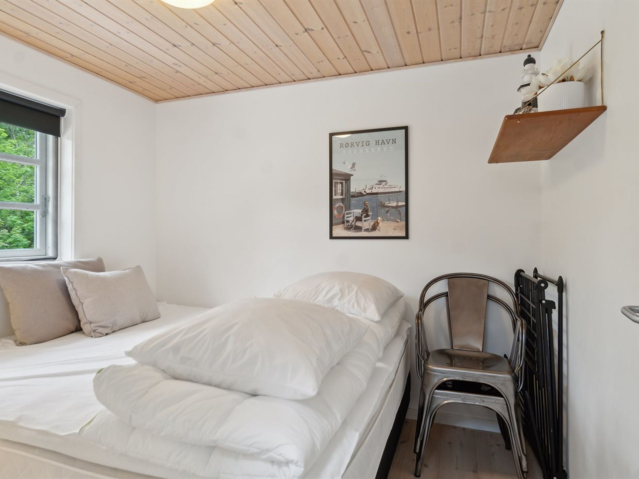 Ein Schlafzimmer mit einem Bett, Kissen, einem Sessel, Holzboden, einem Fenster und einem gerahmten Bild an der Wand.