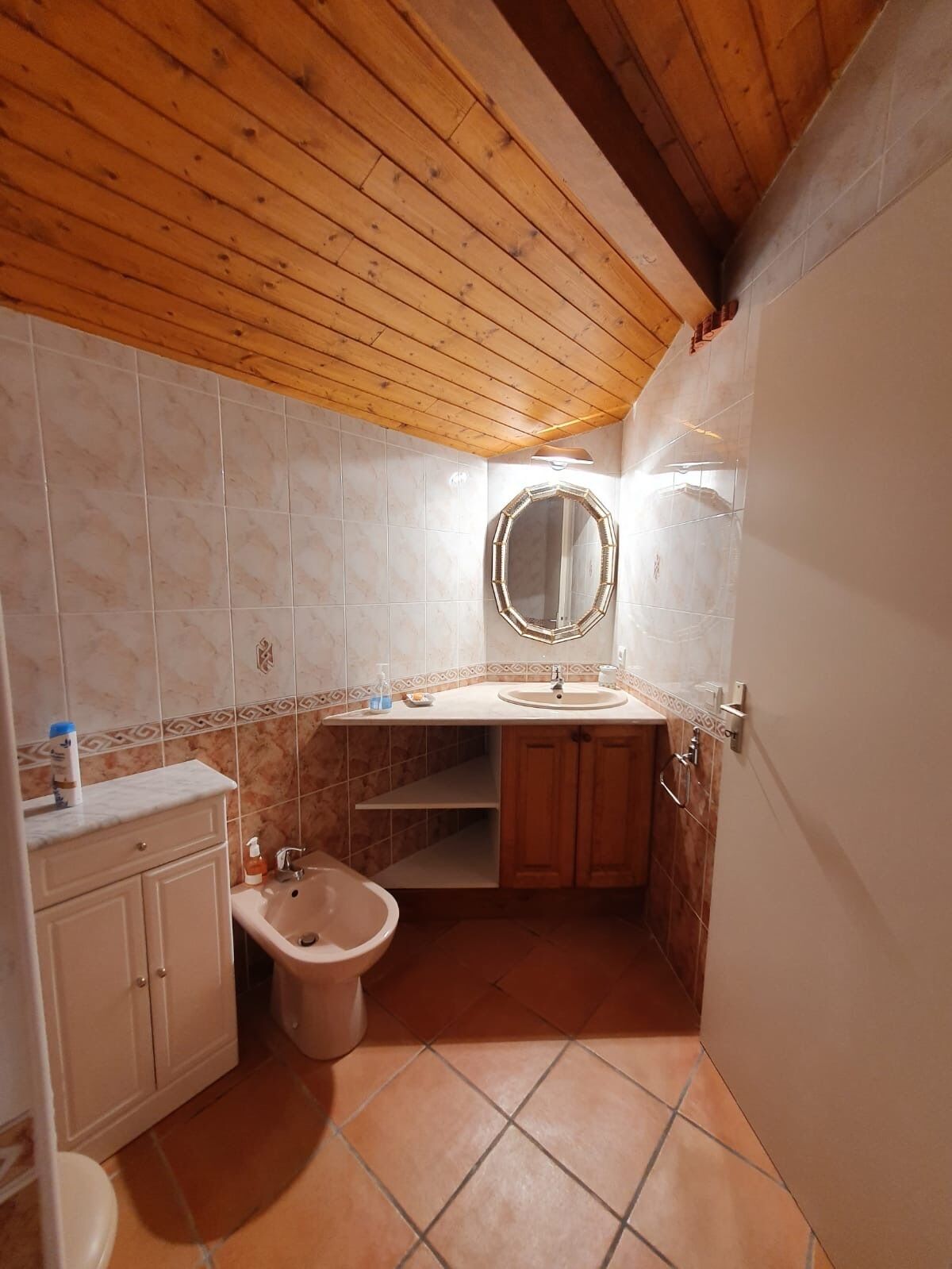 A bathroom with a wooden ceiling, a round mirror, a sink, a bidet, and tiled walls.