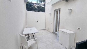 Un espai exterior petit i tancat amb una taula i cadires blanques, un armari blanc i una porta corredissa de vidre.