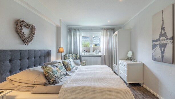Ein Schlafzimmer mit einem großen Bett, einem Schminktisch, einem Nachttisch, einer herzförmigen Dekoration und einem Fenster mit Vorhängen.