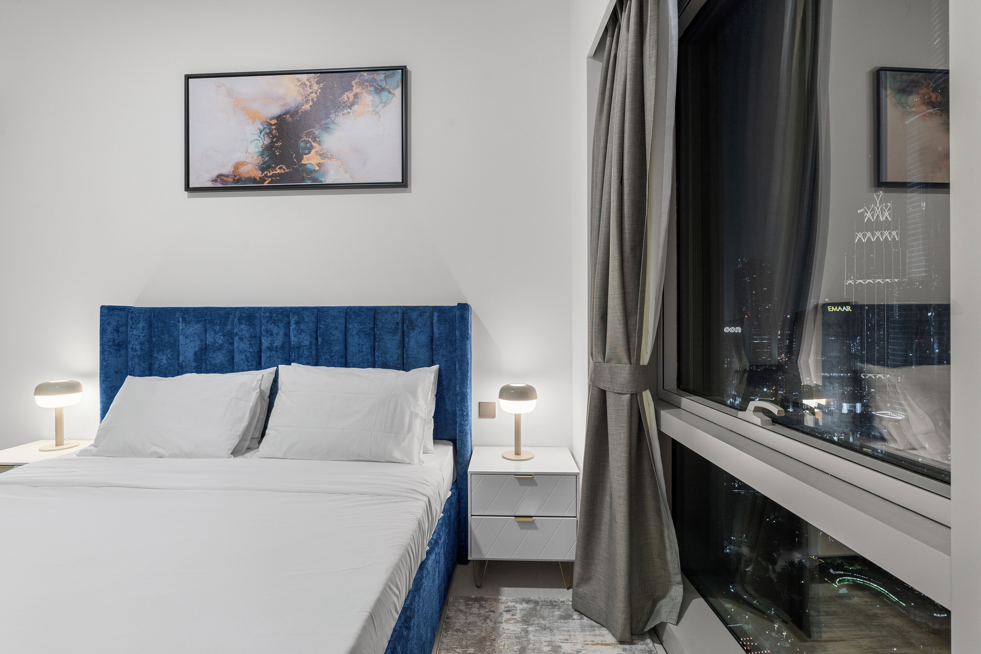 Ein modernes Schlafzimmer mit blauem Kopfteil, weißer Bettwäsche, Blick auf die Stadt durch ein Fenster und einem gerahmten abstrakten Gemälde an der Wand.