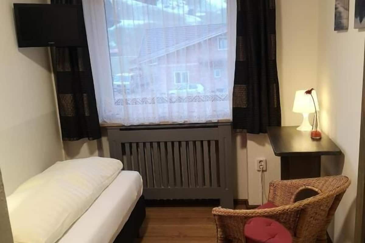 A small, single-room hotel room with a bed, a chair, a table with a lamp, a window with curtains, and a radiator.