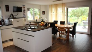 Une cuisine moderne dotée d’un îlot central, d’une table à manger et de chaises. Un téléviseur est fixé au mur et une fenêtre offre une vue sur l’extérieur.