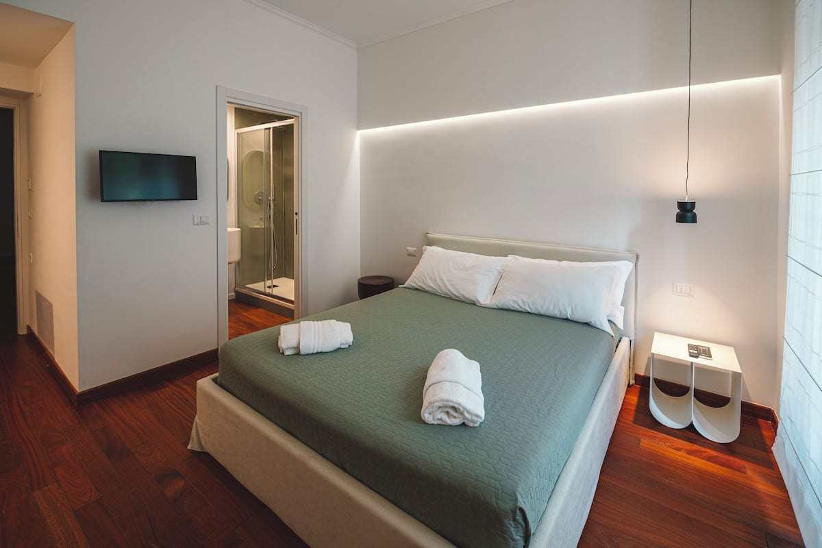 A neatly made bed with a green bedspread, a flat-screen TV mounted on the wall, and a modern bedside table with a lamp.