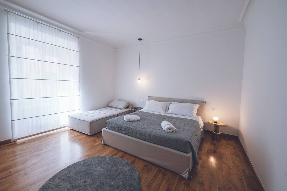 A modern bedroom with a large bed, a smaller sofa bed, a bedside table with a lamp, and a large window with blinds.
