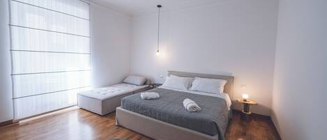 Un dormitorio moderno con una cama grande, un sofá cama más pequeño, una mesita de noche con lámpara y un ventanal con persianas.