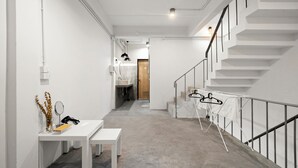 A modern hallway with a white bench, a table with a mirror, and a coat rack. A staircase with a metal railing is visible on the right. - Kuha Phuket Old Town (Phuket)