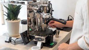 A coffee machine with a digital display, a potted plant, and a metal cup on a counter. - Kuha Phuket Old Town (Phuket)