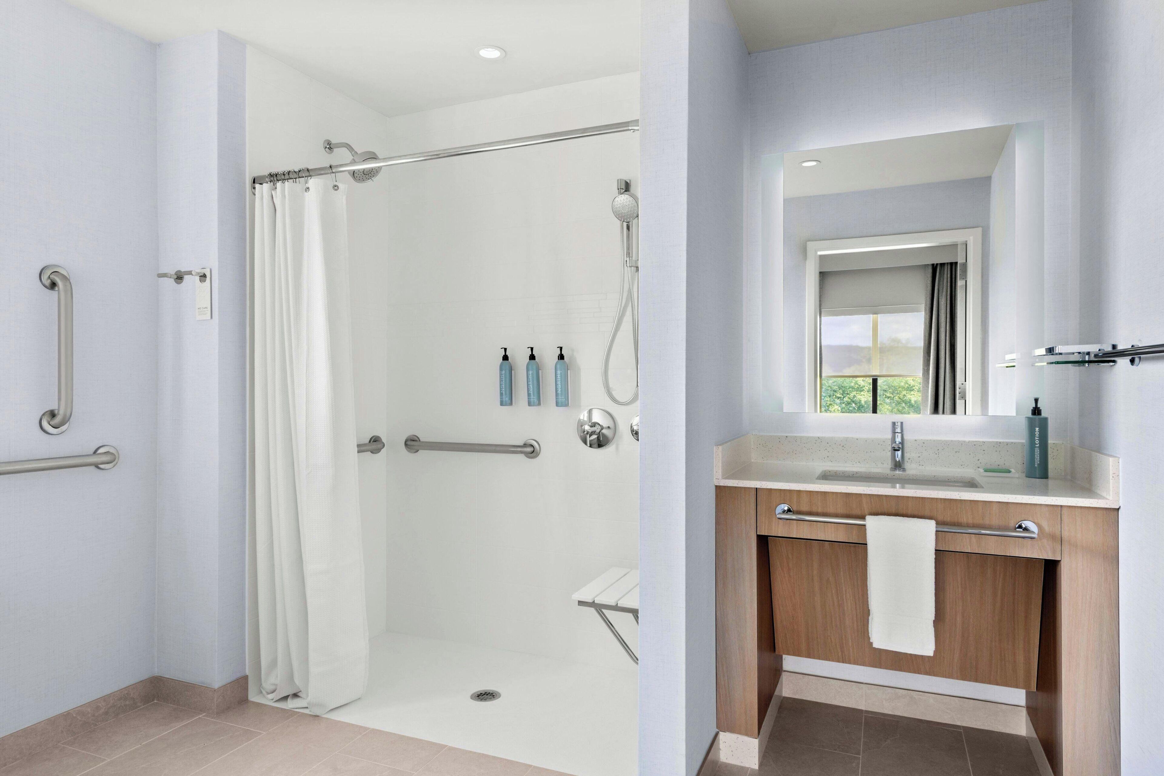 A modern bathroom with a walk-in shower, grab bars, a vanity with a mirror, and a window.