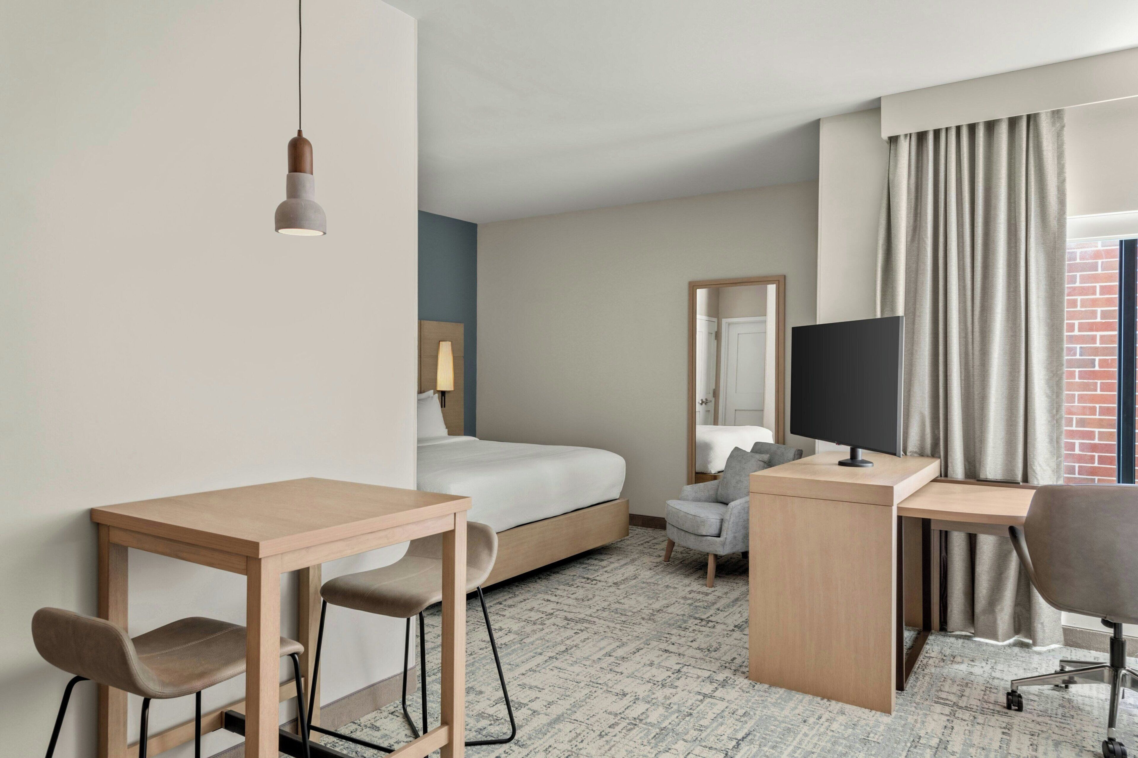 Una habitación de hotel con cama, escritorio, dos sillas, televisor y ventana con cortinas.