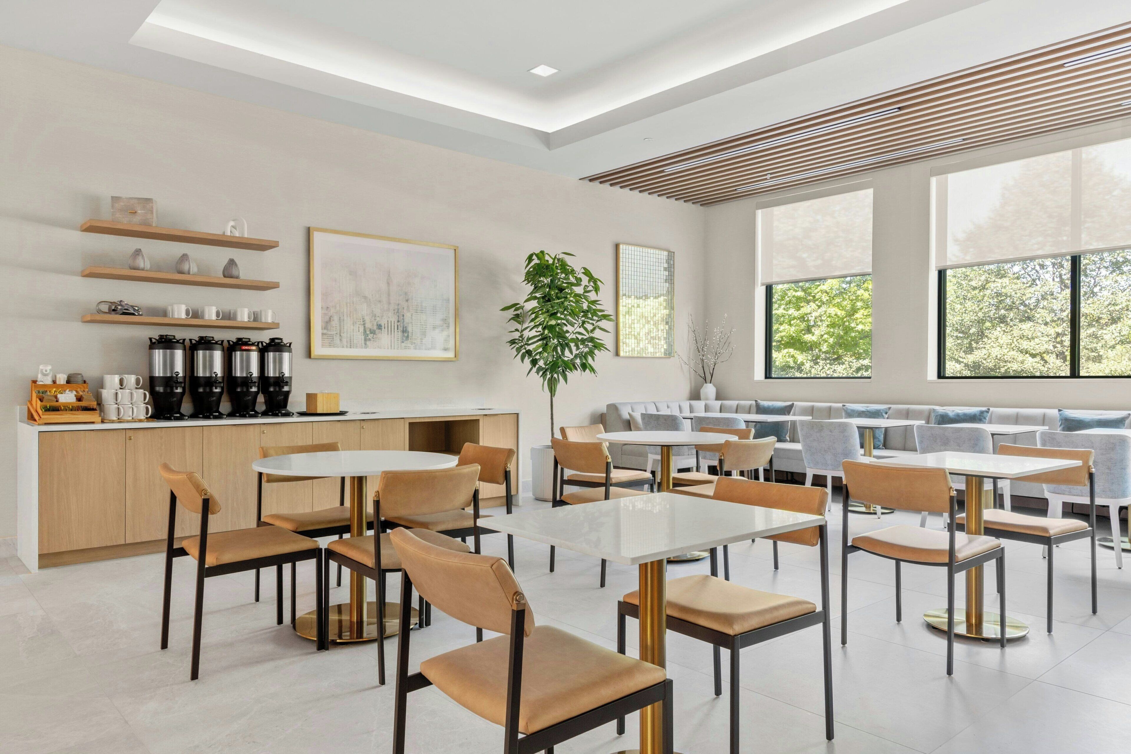 A modern dining area with round tables and chairs, a bar counter with coffee makers, and large windows offering views of greenery.