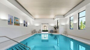Une piscine intérieure moderne, avec une eau d’un bleu cristallin, un plafond en bois et une vue sur un jardin grâce à une grande fenêtre.