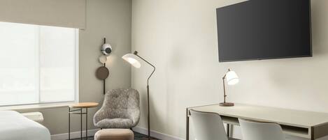 A hotel room with a bed, a desk with two chairs, a lamp, a round table, and a wall-mounted TV.