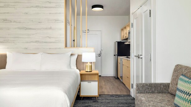 Ein modernes Hotelzimmer mit Bett, Nachttisch mit Lampe, einer kleinen Sitzecke, einer Küchenzeile und einer Tür zu einem weiteren Raum.