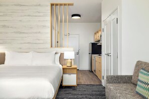 Una habitación de hotel moderna con cama, mesita de noche con lámpara, un pequeño rincón de descanso, kitchenette y una puerta que da a otra habitación.