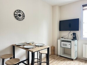 A dining area with a wooden table, chairs, a clock, a TV, and a microwave on a cart.