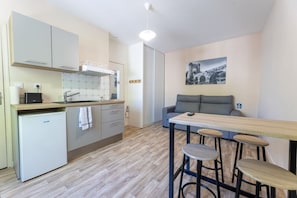A compact kitchen with a refrigerator, a sink, and a stove. There is a dining area with a table and bar stools. A sofa is placed against the wall.
