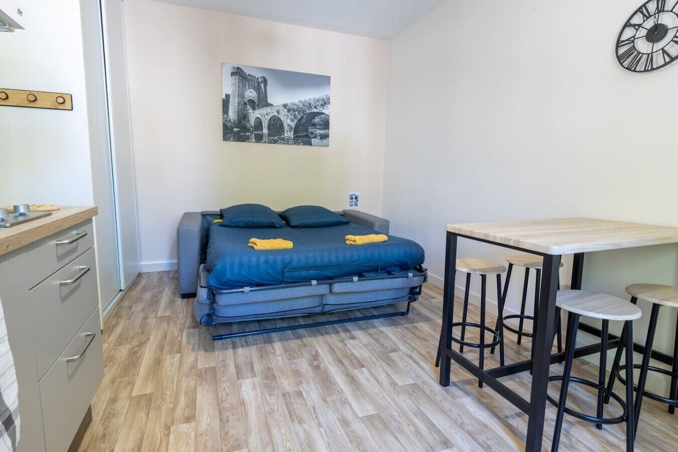 Habitación pequeña con cama, una mesita, un taburete, un reloj en la pared y una imagen de un puente.