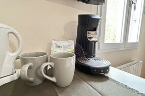 Une machine à café, deux tasses et un paquet de café sont posés sur une table près d’une fenêtre.