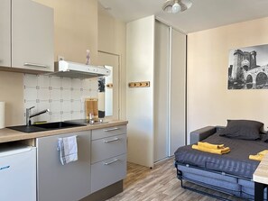 A compact room with a kitchenette, a bed with a dark gray mattress, and a painting of a historical building on the wall.