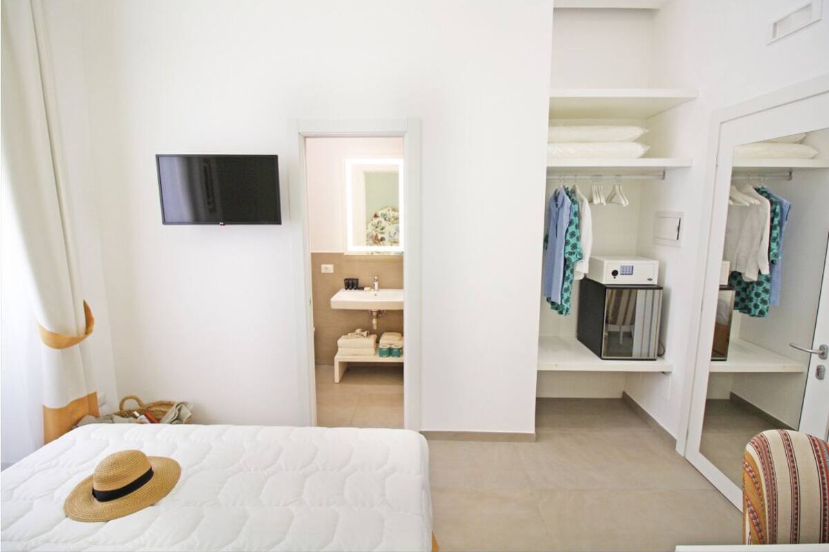 A hotel room with a bed, a TV mounted on the wall, a bathroom with a sink and mirror, and a wardrobe with hanging clothes.