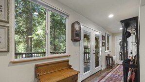 A hallway with a wooden bench, a large window with a view of trees, and a black cabinet with a mirror. - Nana’s Cozy Cottage in Long Barn – Gated Mountain Retreat (Mi-Wuk Village)