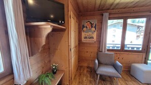 Interior de una cabaña de madera con un televisor montado en la pared, un cartel, una silla, una mesita con una planta y una ventana con cortinas.