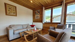 Una sala de estar acogedora con techo de madera, un sofá gris, dos sillones, una mesa de centro de madera y un cartel en la pared.