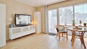 Una sala de estar moderna con un televisor de pantalla plana sobre un mueble blanco, una mesa de comedor con sillas y ventanas amplias con cortinas translúcidas.