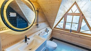 A small, wooden cabin bathroom with a round mirror, a sink, a toilet, and a window.