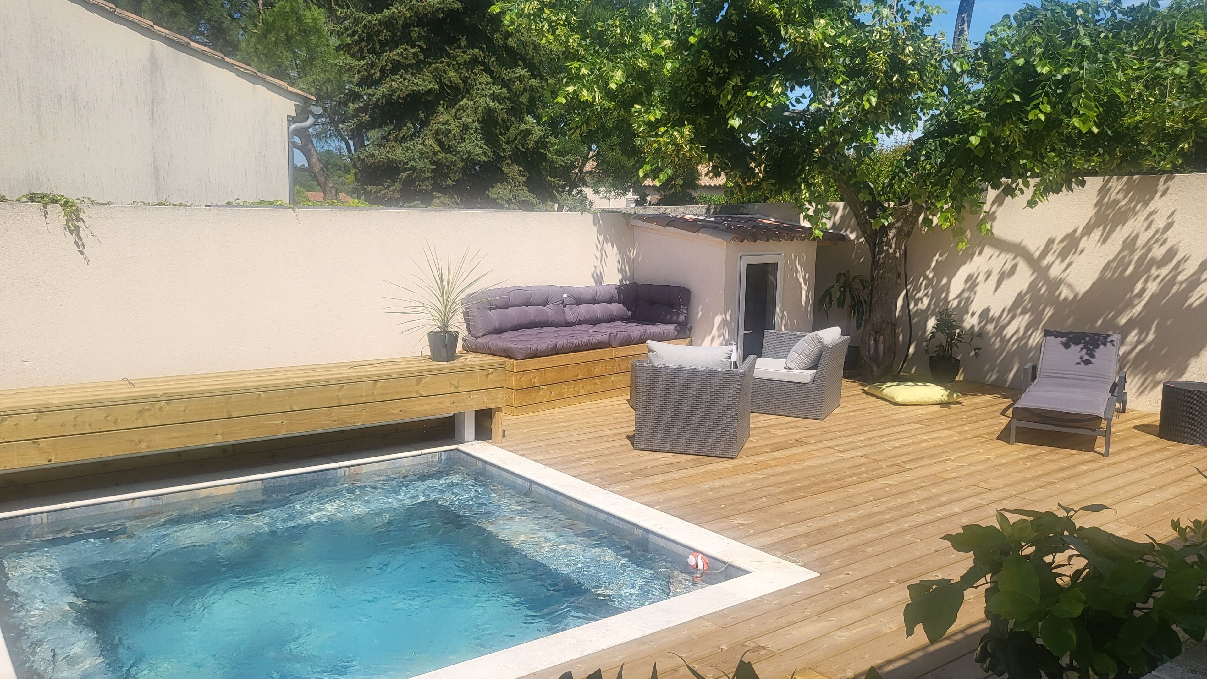 Un patio trasero con piscina, terraza de madera y zona de estar exterior con cojines y una silla.