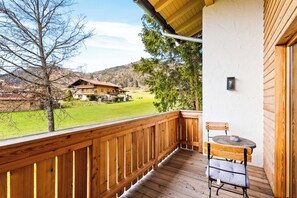 Um deck de madeira com uma mesa e uma cadeira, com vista para uma paisagem verde e um edifício ao longe.