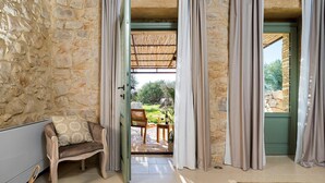 Une pièce aux murs de pierre, avec un canapé et des rideaux, donnant sur une terrasse extérieure équipée d’une table et de chaises.