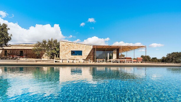 Un espace aménagé au bord de la piscine, avec un bâtiment en pierre, des sièges d’extérieur et un ciel d’un bleu limpide.