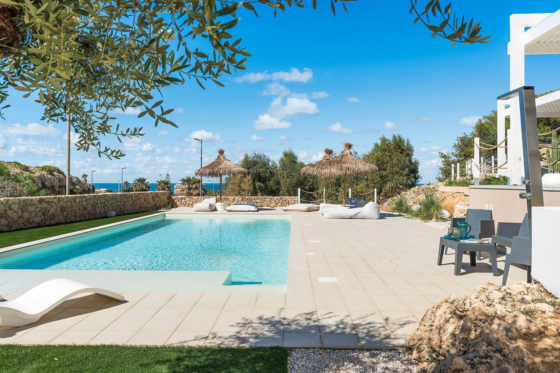 A swimming pool with lounge chairs and a thatched umbrella, surrounded by a stone wall and greenery, with a view of the sea.