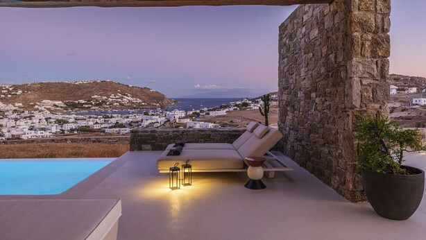 A modern outdoor lounge area with a pool, a stone wall, and a view of a coastal town.
