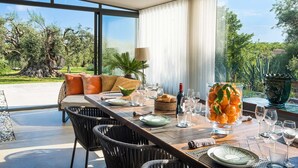 Un comedor con una mesa larga dispuesta para seis personas, rodeado de sillas de mimbre, un sofá con cojines y vistas a un jardín a través de grandes ventanales.