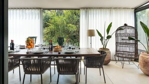 Un comedor con una mesa dispuesta para cuatro personas, rodeado de sillas de mimbre, y vistas a la vegetación a través de amplios ventanales.