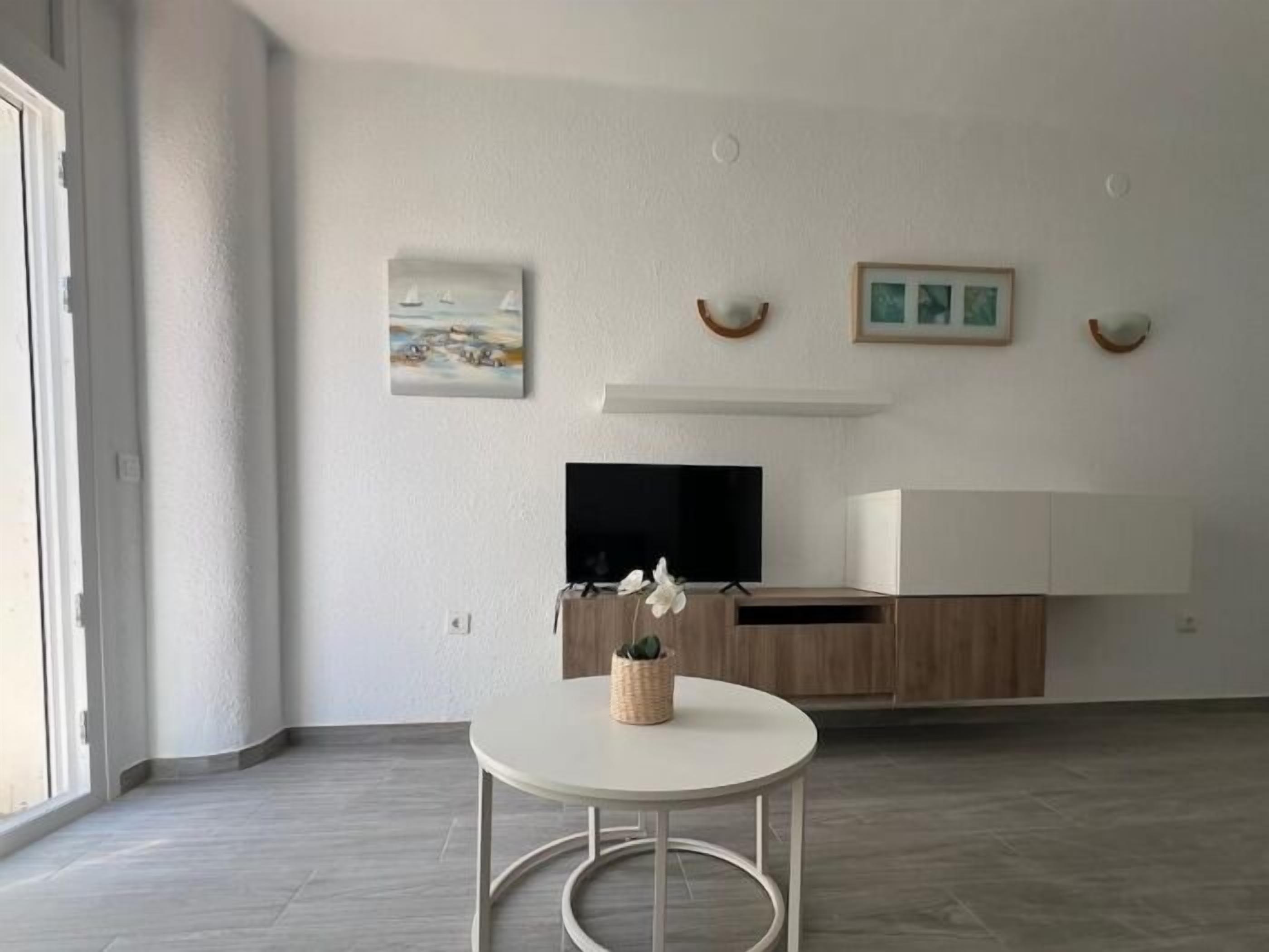 A modern living room with a flat-screen TV, a small round table with a plant, and artwork on the wall.