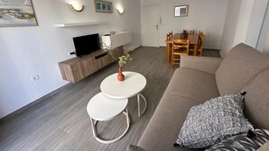 Ein modernes Wohnzimmer mit einer Couch, einem kleinen weißen Tisch, einem Fernseher auf einer Holzkommode und einem Essbereich mit Holztisch und Stühlen.