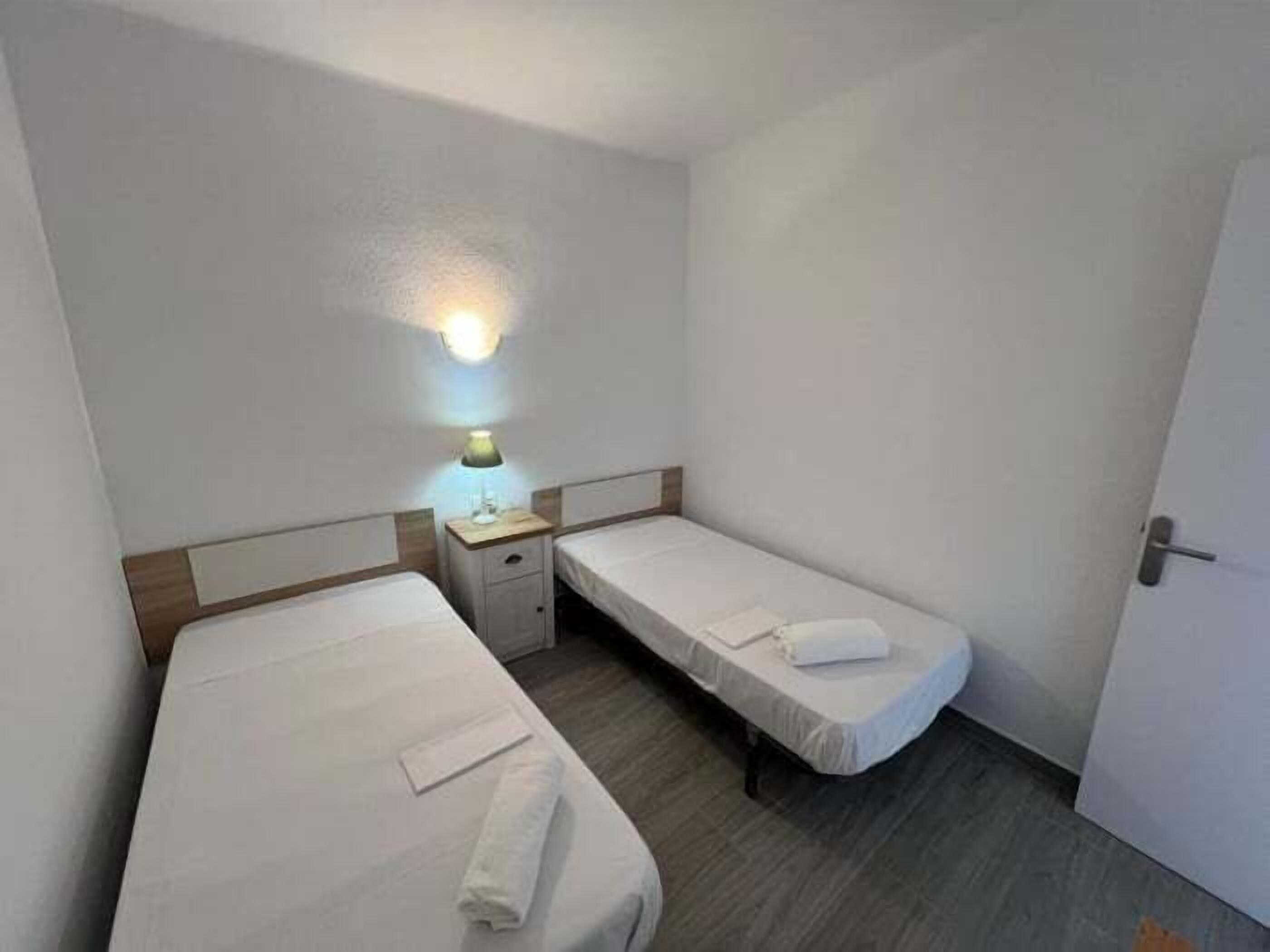 A small, clean room with two single beds, a nightstand with a lamp, and a door.