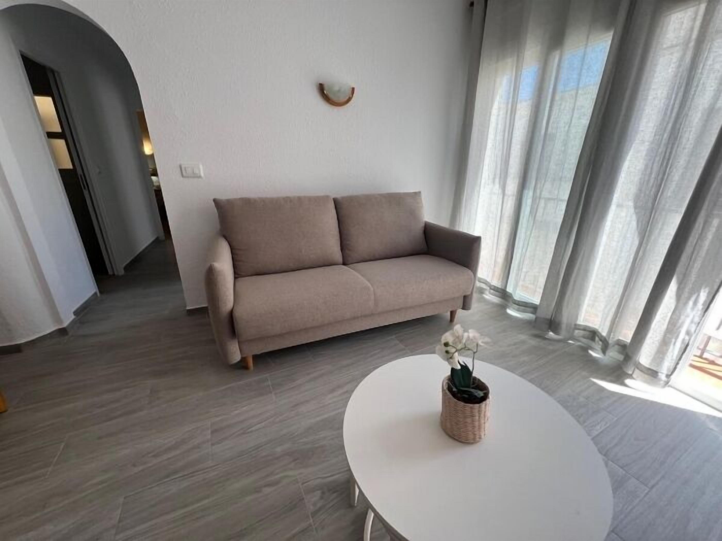 A modern living room with a beige sofa, a round white table with a potted plant, and a large window with sheer curtains.