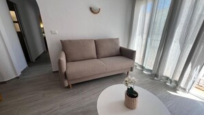 Ein modernes Wohnzimmer mit einem beigefarbenen Sofa, einem runden weißen Tisch mit einem Blumentopf und einem großen Fenster mit transparenten Vorhängen.