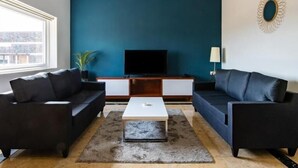 A modern living room with a dark blue accent wall, a ceiling fan, a flat-screen TV, and two black sofas.
